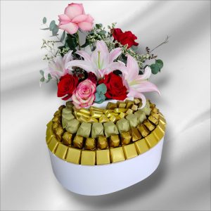 "Luxury white oval gift box with a tiered arrangement of gold-wrapped chocolates, topped with a fresh bouquet of pink roses and white lilies.