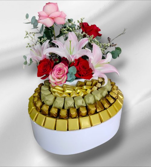 "Luxury white oval gift box with a tiered arrangement of gold-wrapped chocolates, topped with a fresh bouquet of pink roses and white lilies.